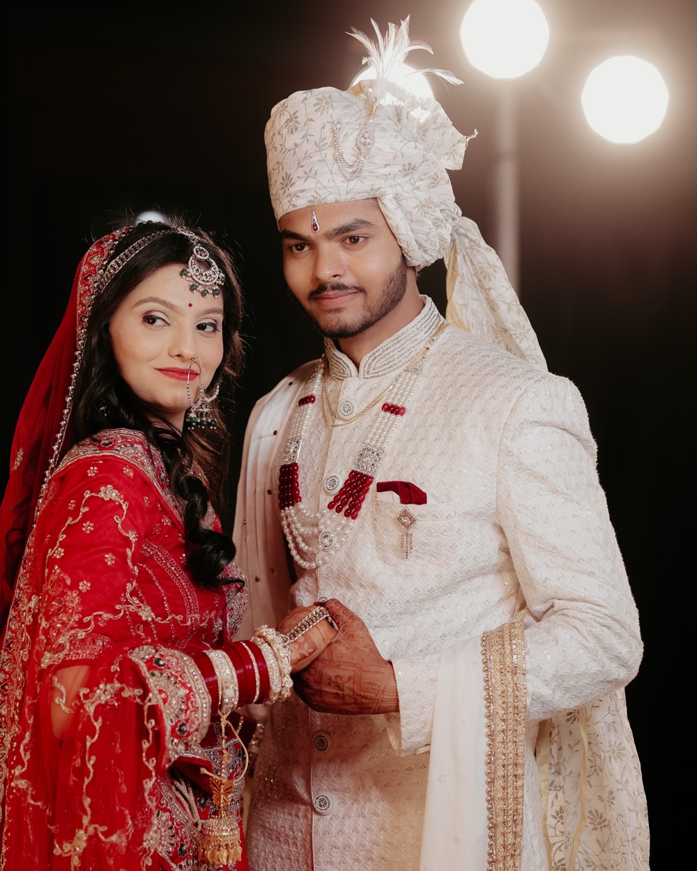 Bride in red and groom in an ivory feathered turban standing together under stage lights