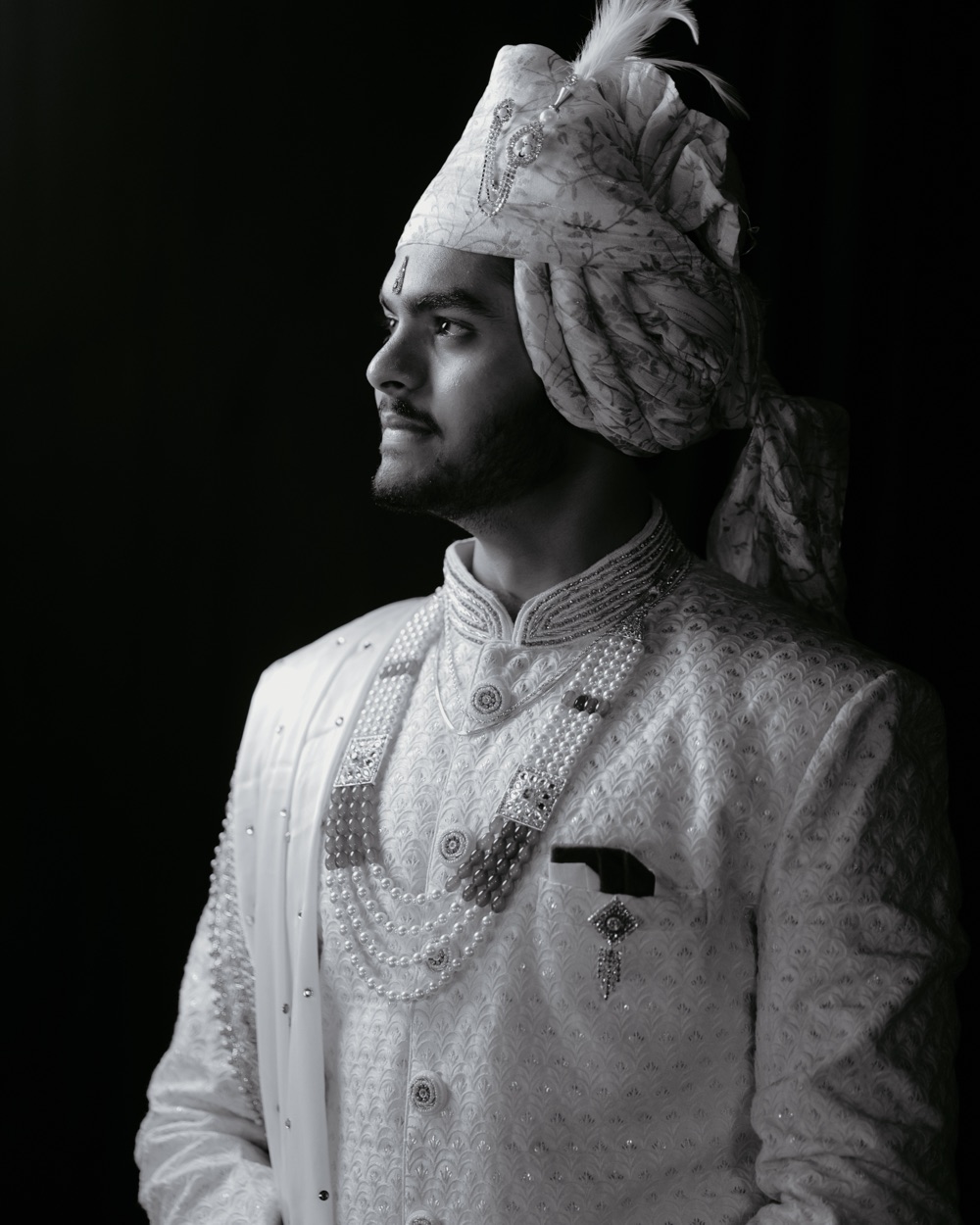 Black-and-white profile portrait of the groom in an ivory sherwani and feathered turban