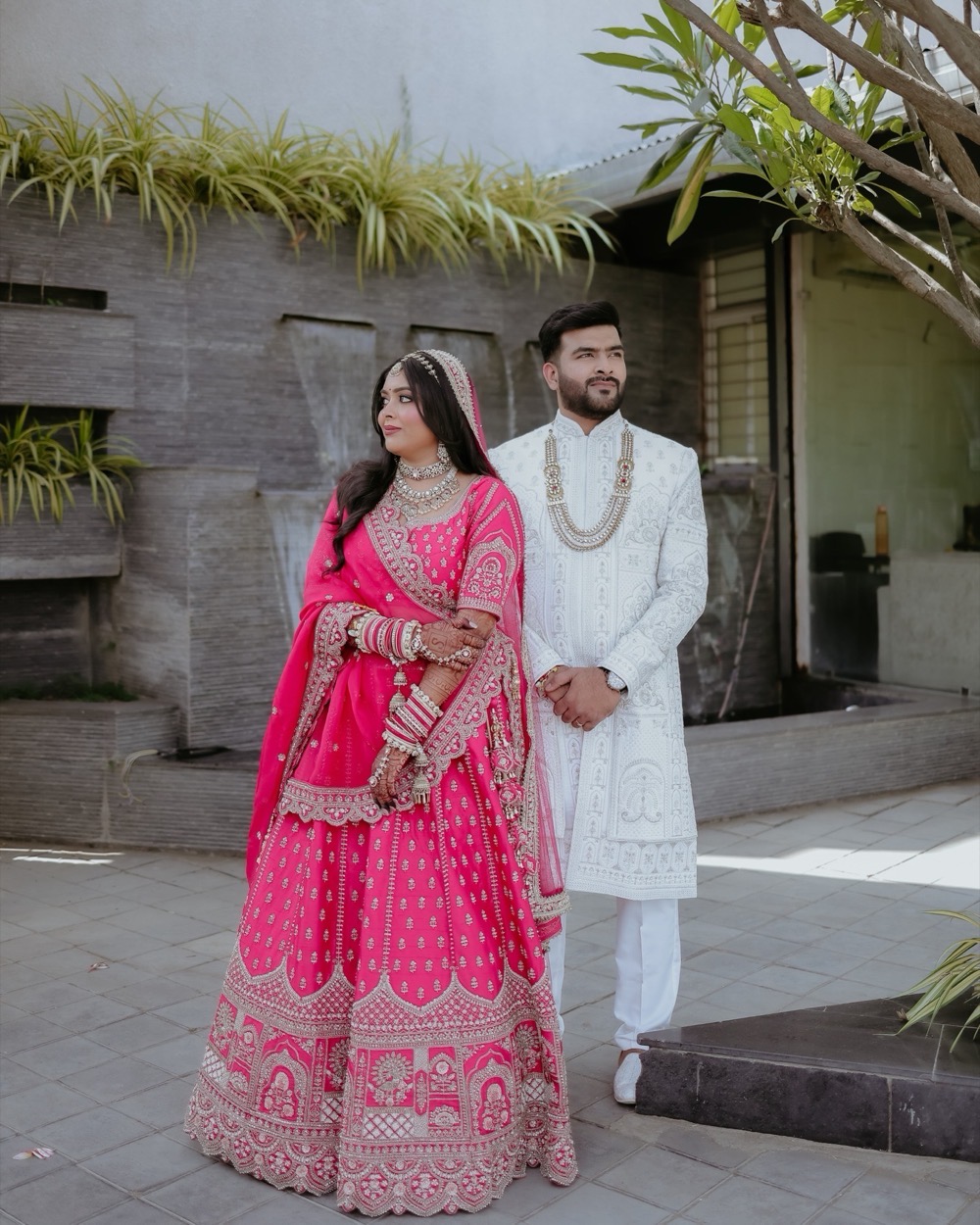 Bride in a rani-pink lehenga and groom in an ivory sherwani standing together in a sunlit courtyard