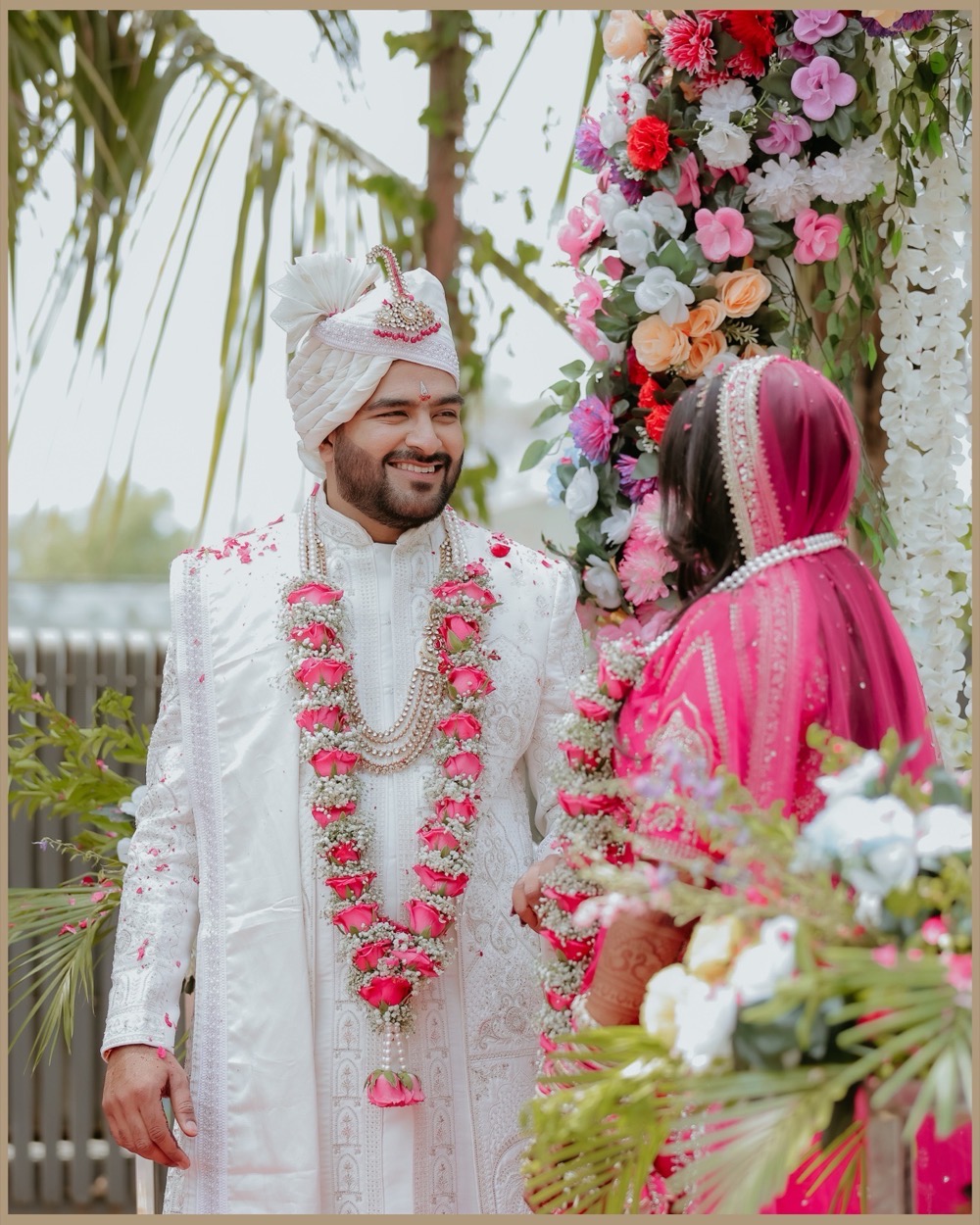 Groom in an ivory sherwani and rose garland smiling at the bride during the varmala ceremony