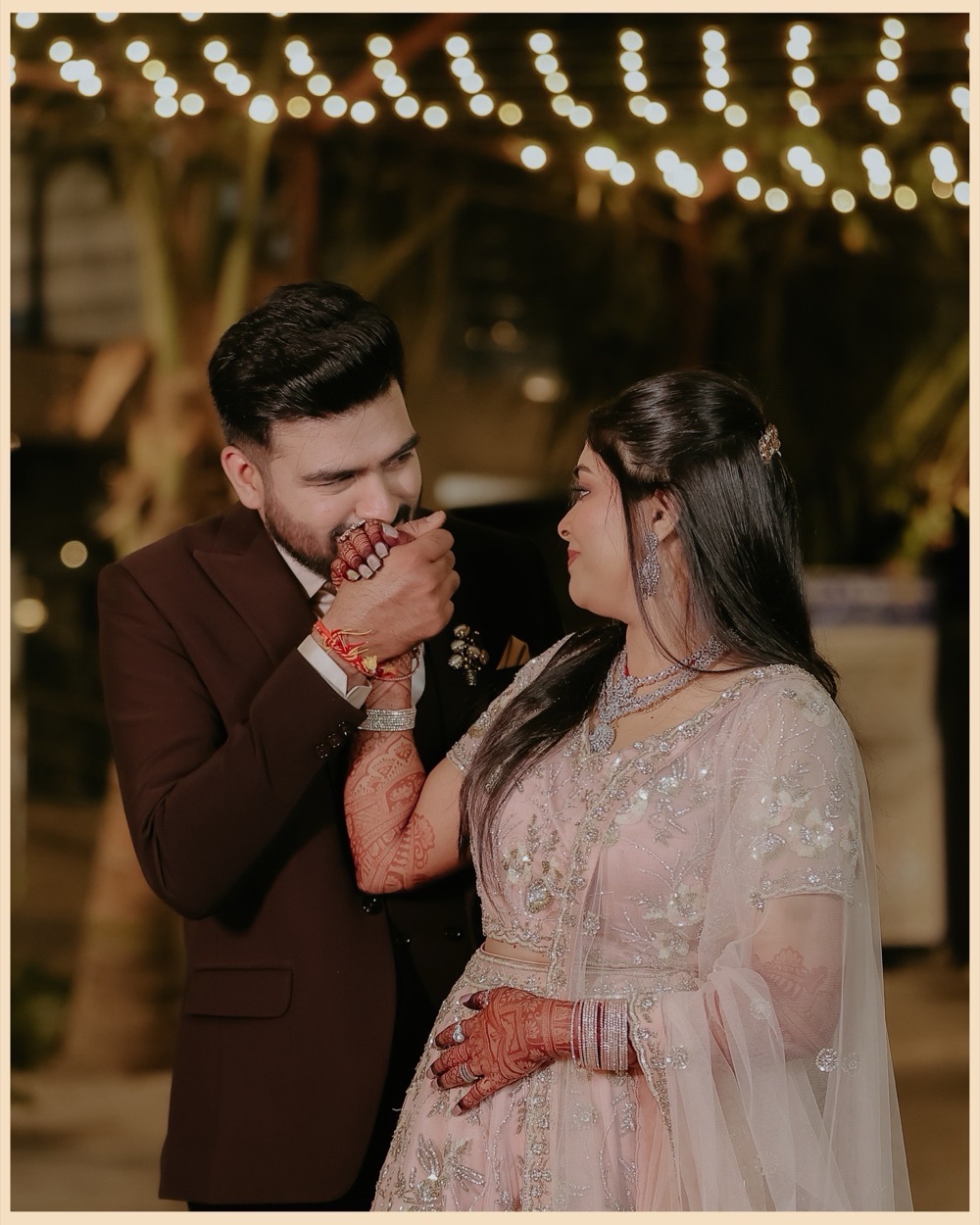 The groom kisses the bride's hennaed hand beneath strings of fairy lights
